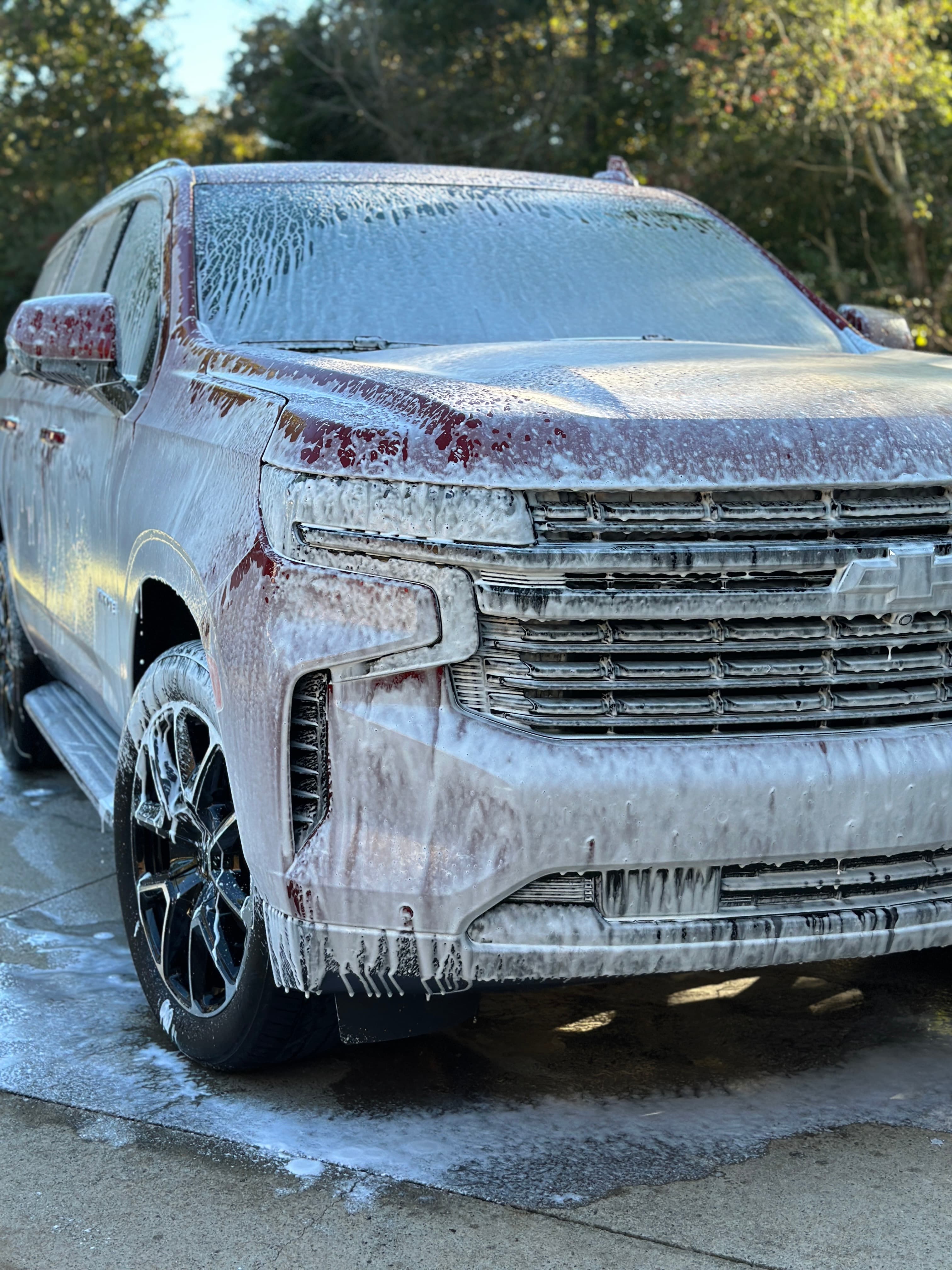 Red SUV covered in thick white soap foam during a car wash outdoors.