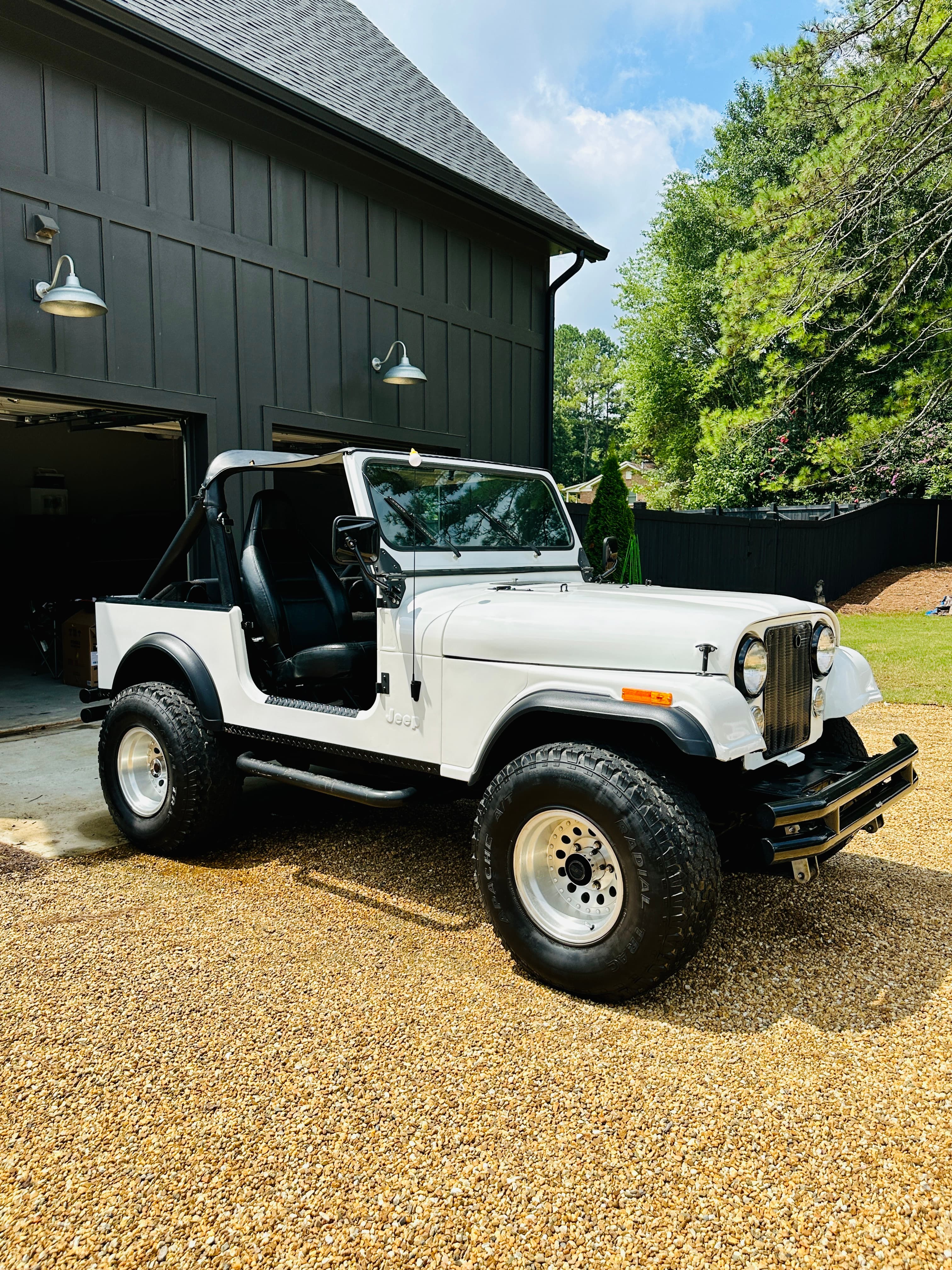 White vintage Jeep with off-road tires parked on a gravel driveway beside a black barn.