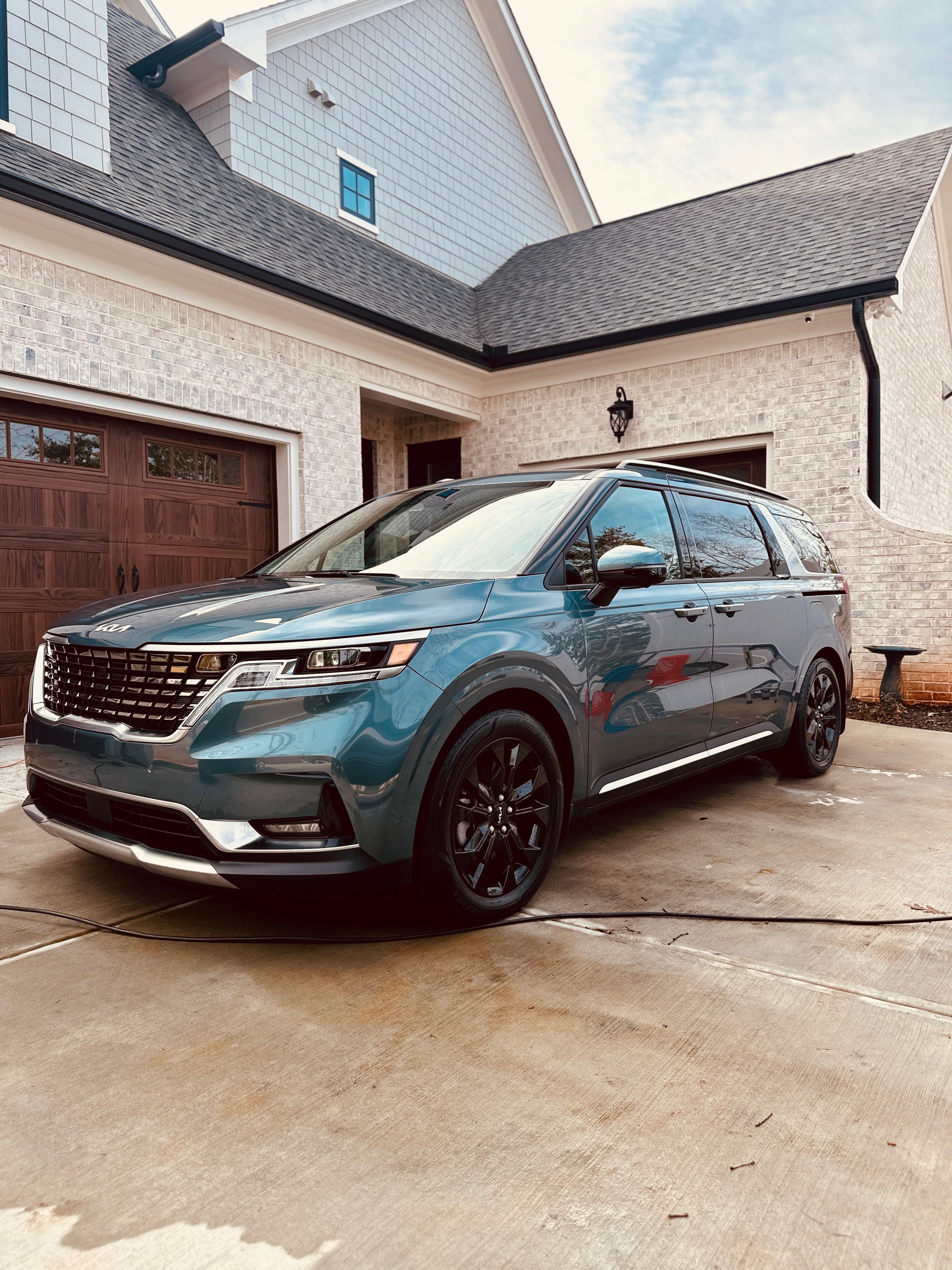 Teal Kia Carnival minivan with black wheels parked on a driveway before a modern house.