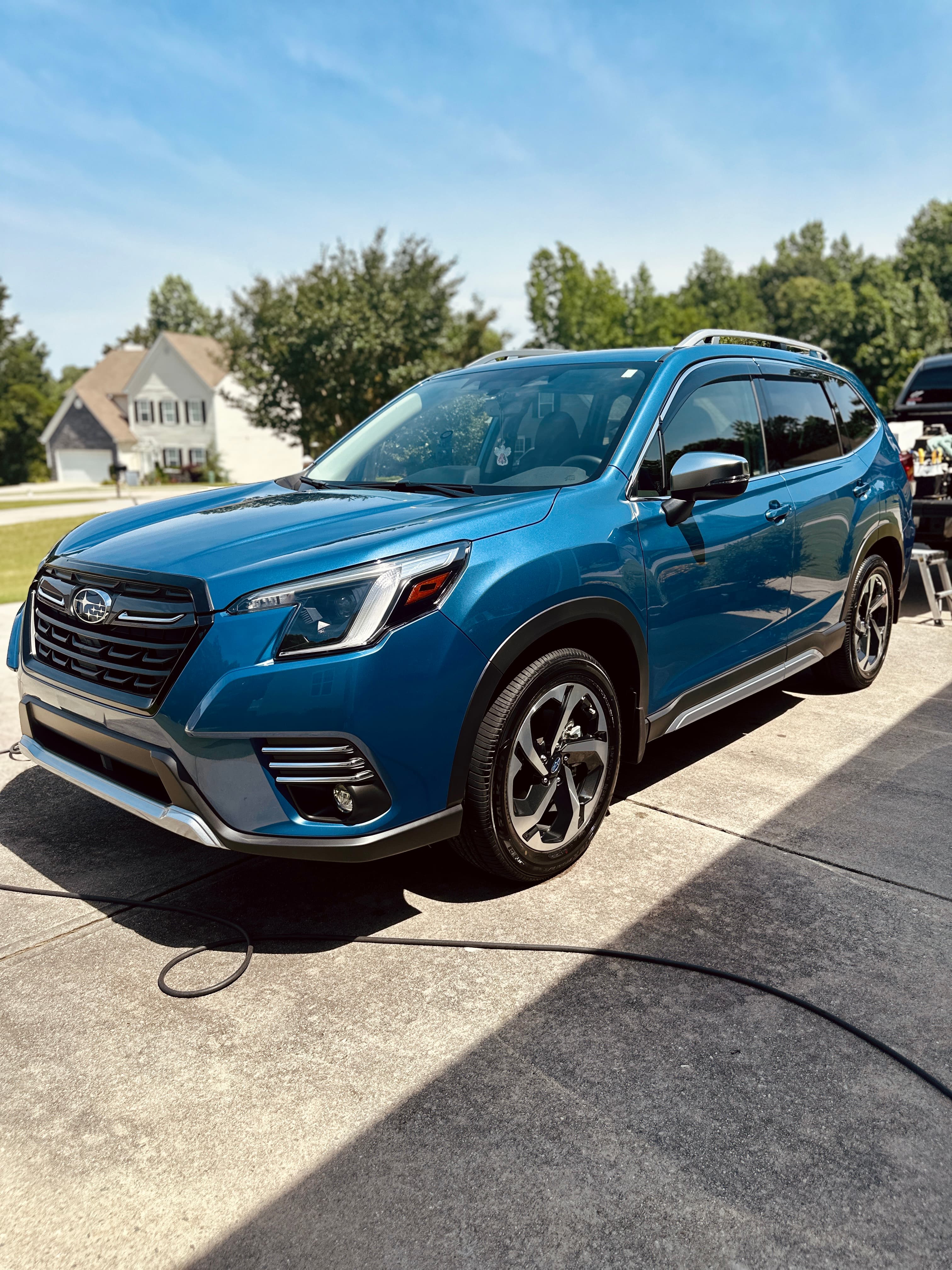 Blue Subaru Forester SUV parked on a concrete driveway in a sunny suburban neighborhood.